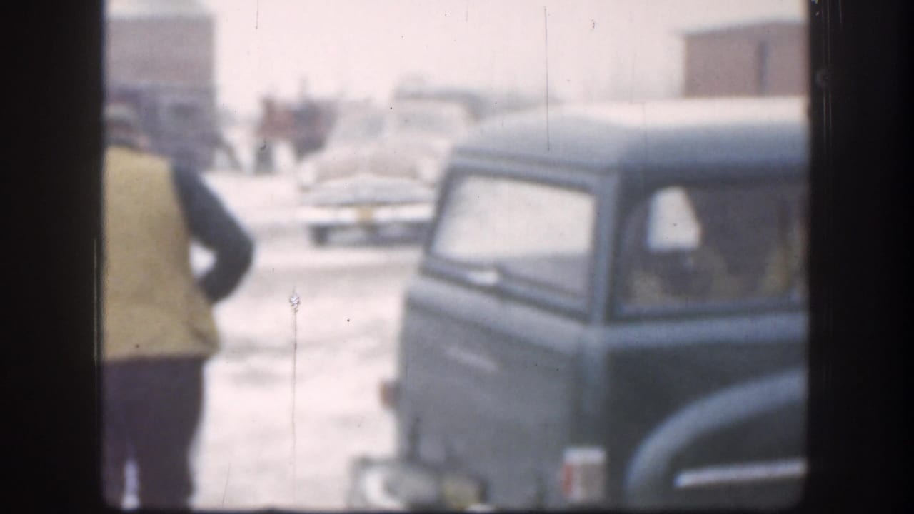 1958 Kansas home-movie frame showing a bundled adult near a dark mid-century car in snowy, low-visibility conditions.