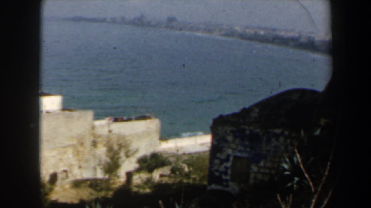 White minaret and stone buildings beside blue sea, partly blocked by dark film-frame edges.