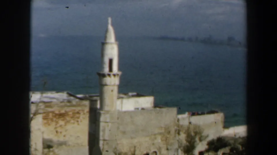 1960 Israel 8mm frame showing a minaret, flat-roofed coastal buildings, and blue water beyond.