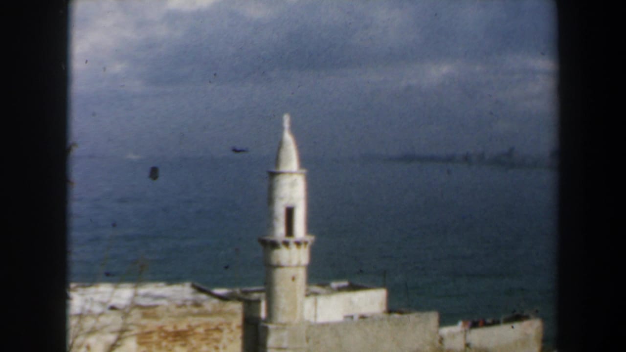 1960 8mm view in Israel of a white minaret rising above coastal buildings, with calm water and clouds in the background.