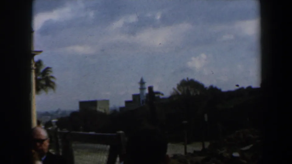 Visitor-shot 1960 Israel coastal view framed by a dark edge, with palm trees, distant buildings, and a man in glasses partly visible.