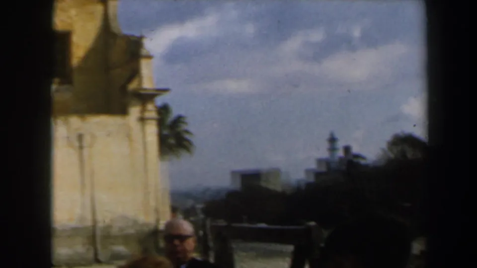 Soft 8mm frame in Israel showing a stone building edge, distant tower shapes, palm foliage, and a man partly visible near the bottom.
