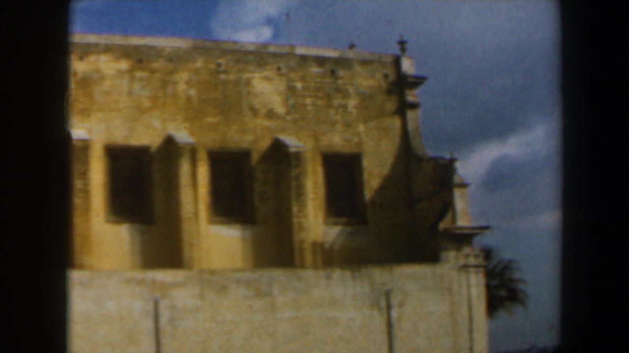 1960 8mm frame from Israel showing a weathered church facade with dark openings, projecting shutters, and blue sky.