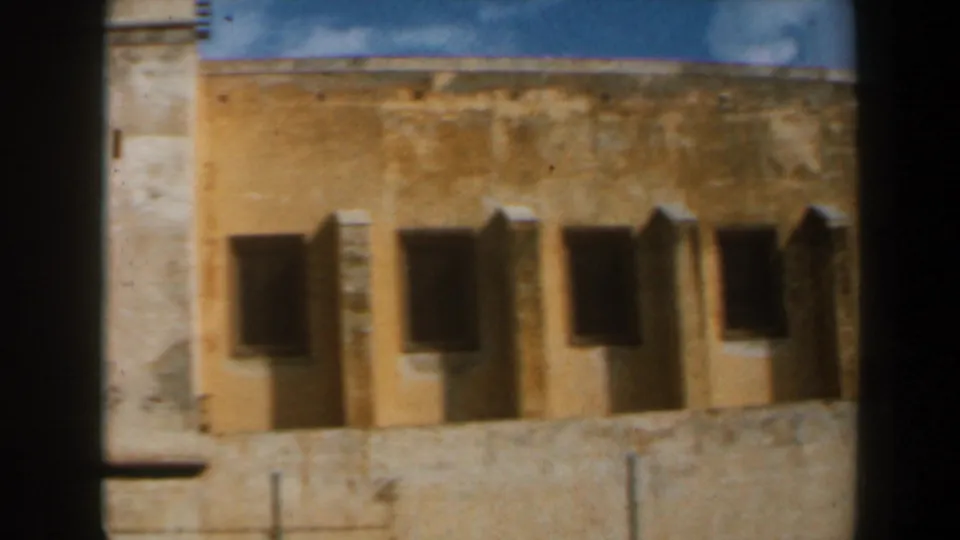 1960 8mm frame from Israel showing a yellowed church wall, shaded window openings, and the edge of a pale bell tower.