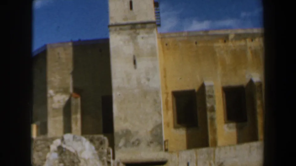 1960 8mm frame from Israel showing a partial church tower, yellow wall, shuttered openings, and rough stonework below.