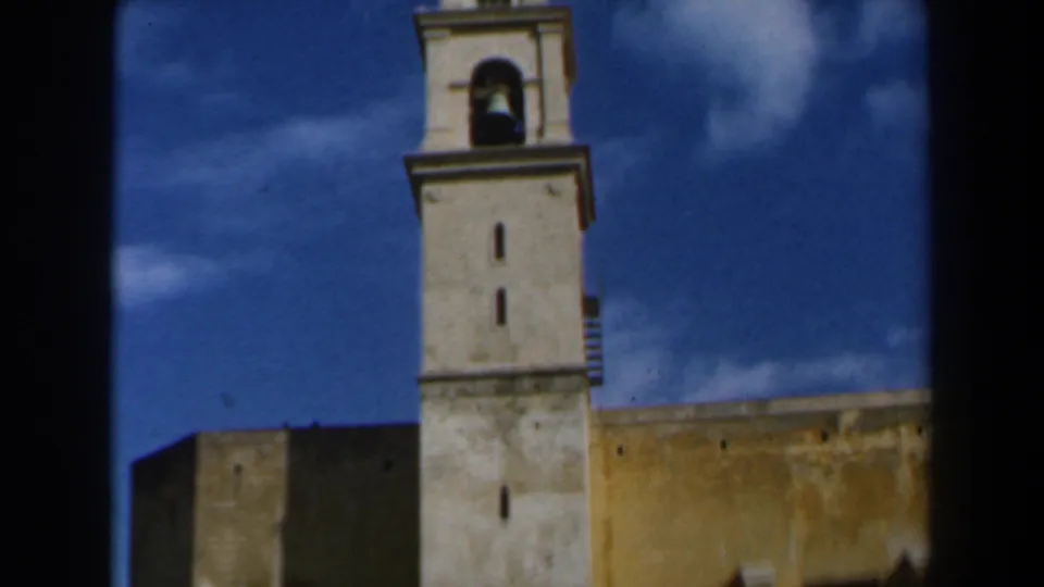 1960 8mm frame from Israel showing a tall church bell tower with a visible bell beside an aged yellow wall.