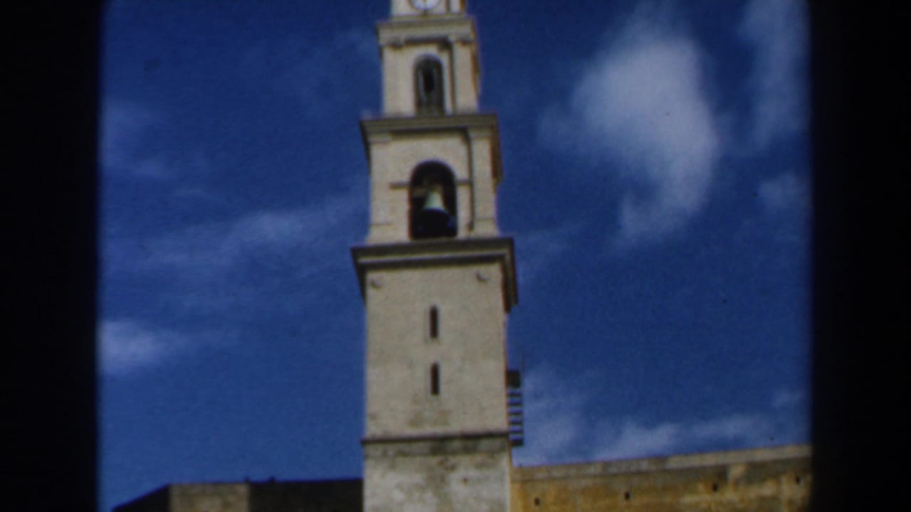 1960s Israel 8mm frame showing a light-colored church tower rising into a deep blue sky with white clouds.