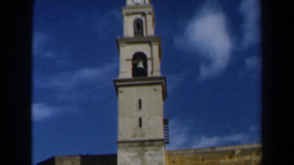 1960s Israel 8mm frame showing a light-colored church tower rising into a deep blue sky with white clouds.