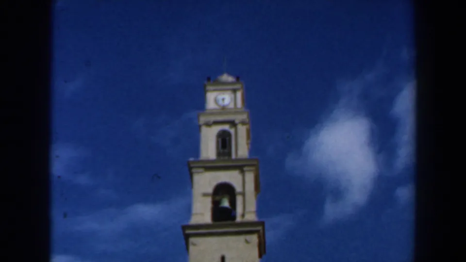 Low-angle 8mm view of the church tower top, showing the bell opening, clock face and saturated blue sky.