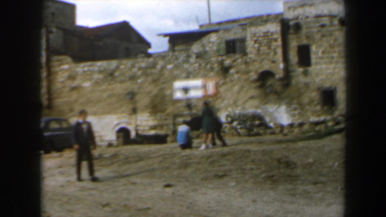 1960s Israel home-movie frame showing a stone courtyard, old masonry buildings, and distant people in daylight.