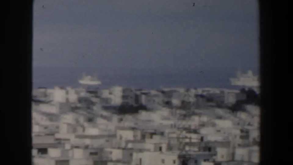 Wide elevated 1960s Israel city view with pale buildings, open sky, sea, and distant ships near the horizon.