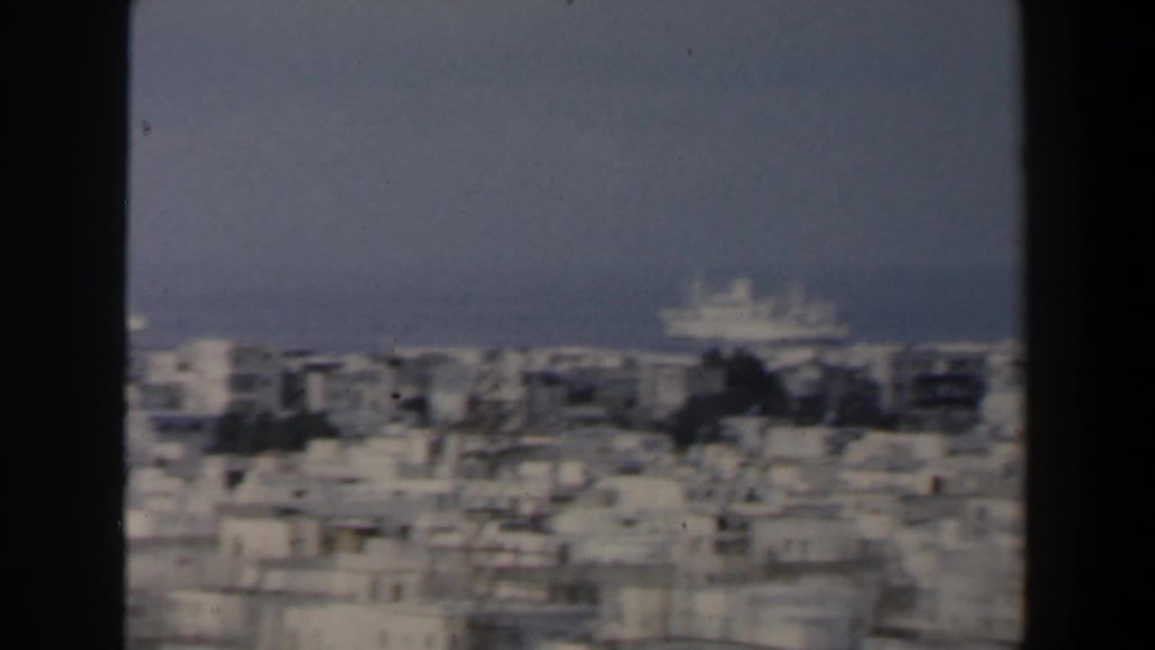 1960 Israel 8mm frame showing a hazy coastal city panorama with white rooftops, sea horizon, and a distant vessel.