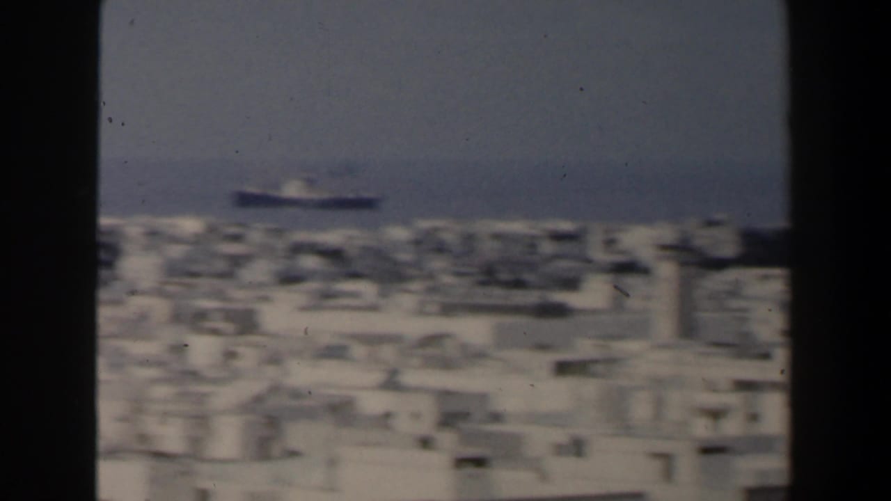 Coastal city rooftops seen through dark vertical framing with a ship offshore.