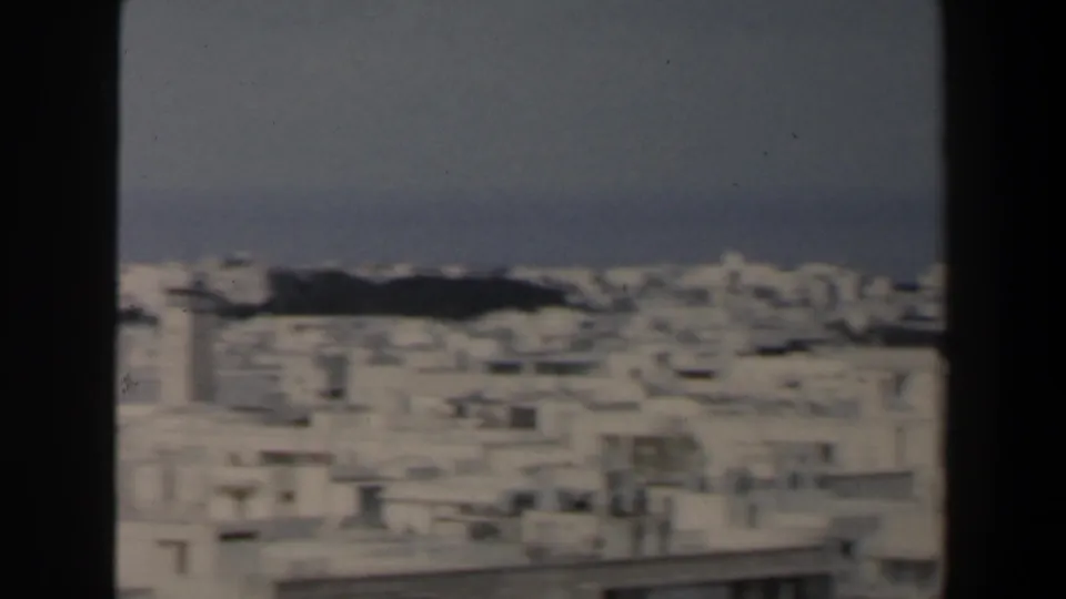 1960 Israel home-movie frame looking across dense white rooftops toward a hazy Mediterranean shoreline.