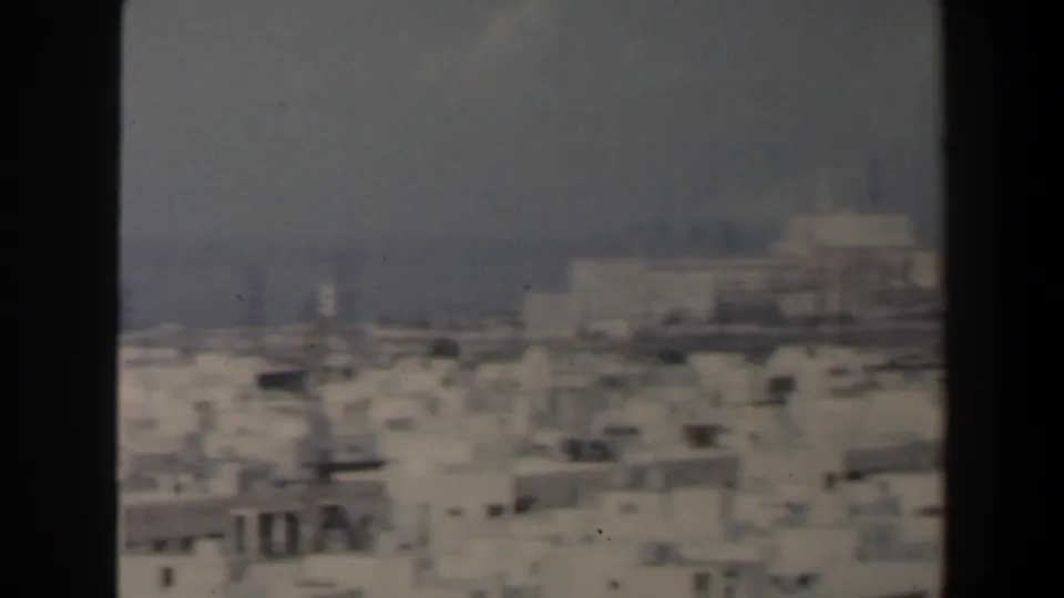 1960 Israel coastal city view with pale buildings in the foreground and a dark ship on the hazy sea.