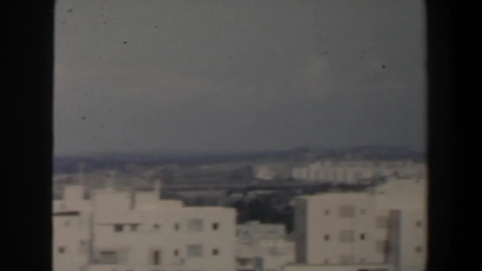 Static 8mm frame of an Israeli cityscape with white rooftops, dark window framing, and a faint distant shoreline under haze.