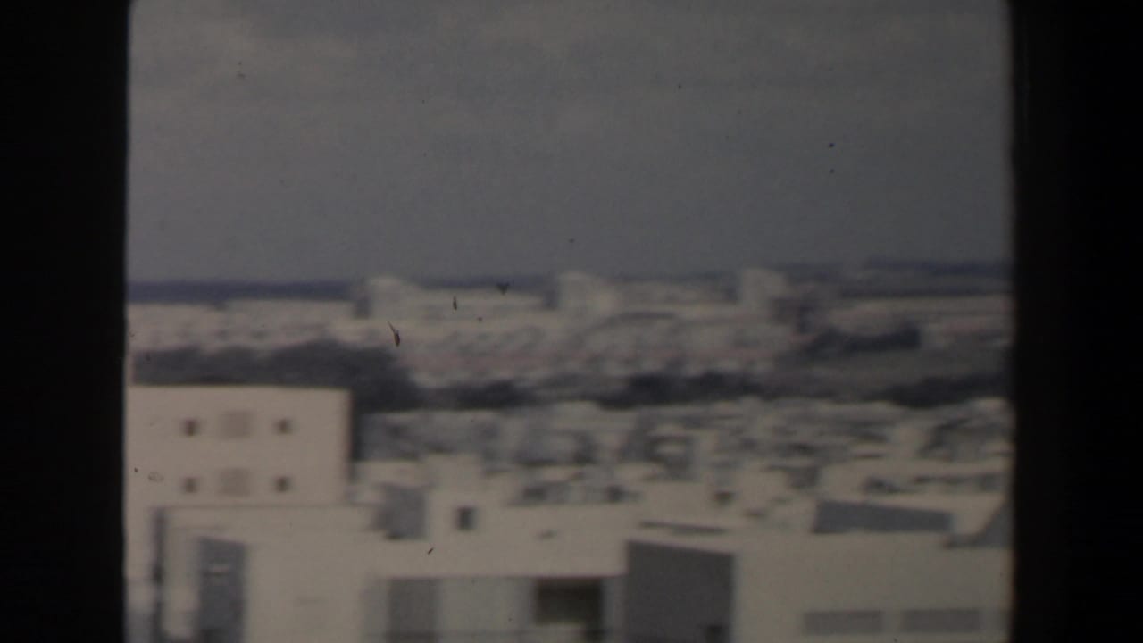 Blurred skyline and white buildings framed by dark film edges.