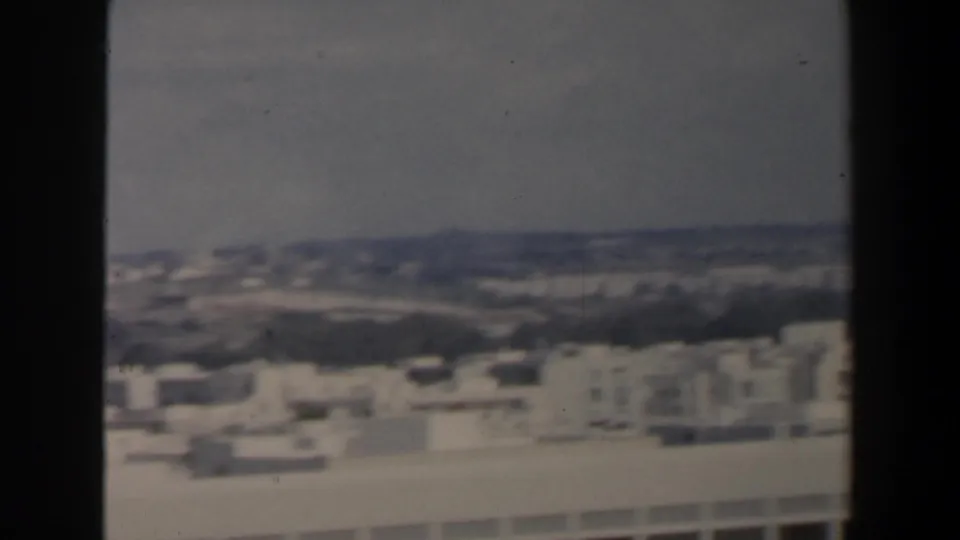 Soft 1960 home-movie panorama across Israeli rooftops toward a distant ridge or shoreline under a pale sky.