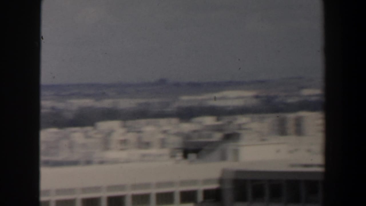 Hazy elevated view across pale low-rise buildings and distant hills, partly bordered by dark vertical frame edges.