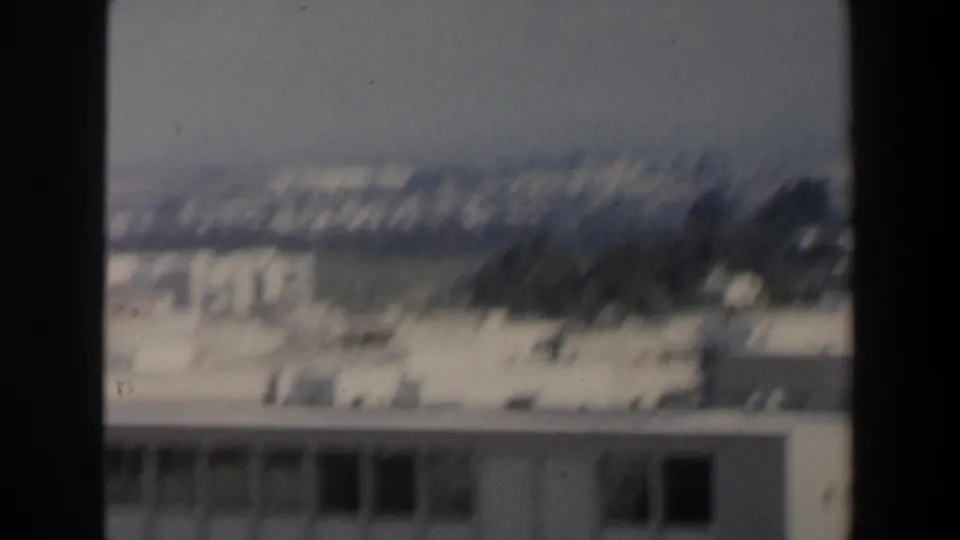 Soft 8mm frame of an Israeli urban view, with distant pale buildings and landscape divided by a dark vertical window or frame edge.