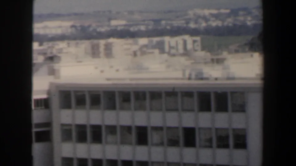 1960 Israel city view with a modern multi-story building in the foreground and pale residential blocks across the hazy background.