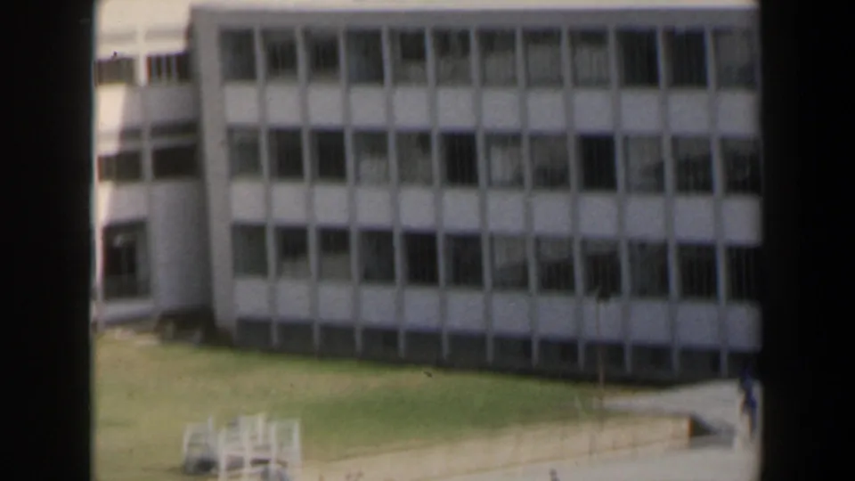 Closer 8mm view of a multi-story building in Israel with repeated window bays, grass below, paths, and distant urban structures.