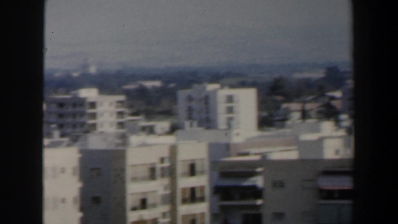 Urban skyline with white high-rise buildings, balconies and distant low hills or trees.