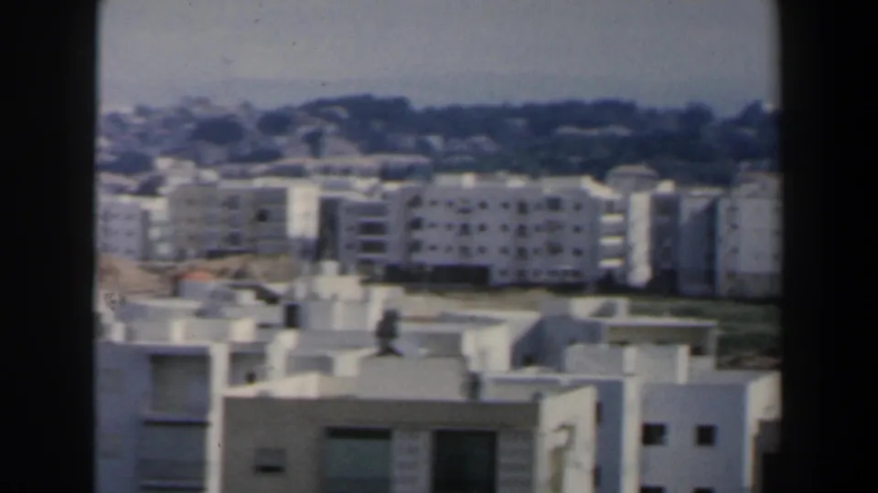 1960 Israel frame showing rows of white residential buildings, roof structures and a distant wooded horizon.