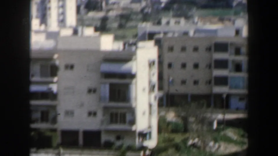 Soft 8mm frame of pale apartment buildings in Israel, with balconies, roofs and blurred greenery in the foreground.