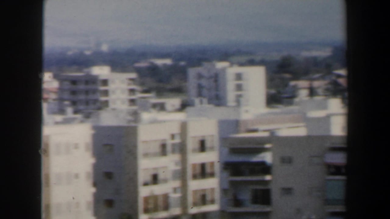 1960 Israel home-movie frame showing pale apartment blocks and distant low-rise city structures under a hazy sky.