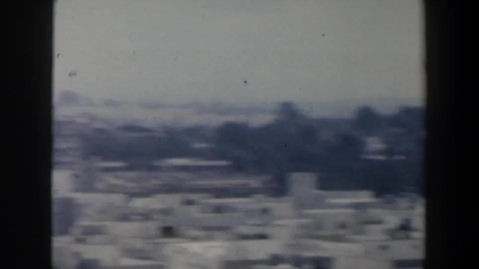 Soft 1960 Israel city-view frame with low rooftops, dark tree shapes, distant buildings, and heavy film softness.