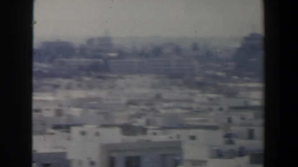 1960 Israel city scan frame with low flat-roofed buildings, distant taller forms, and a central dark vertical obstruction.