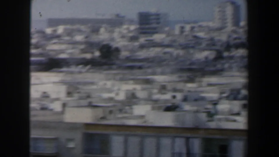 1960 Israel home-movie frame with crowded rooftops, a foreground building facade, and taller distant blocks under bright haze.