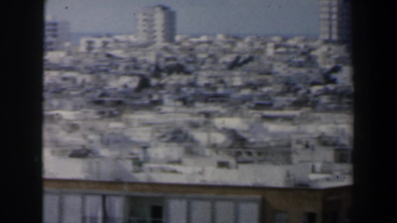 Dense city rooftops with taller buildings rising in the distance.