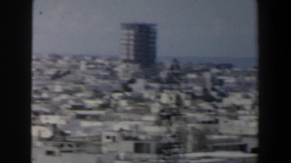 1960s Israel frame showing a hazy city panorama, a prominent high-rise, and a possible distant waterline.