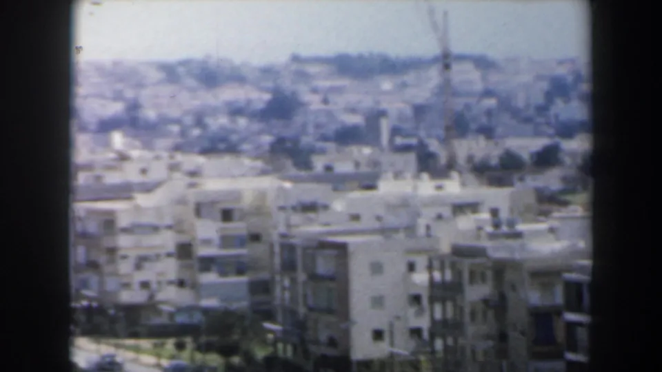Hazy 1960 Israel home-movie frame showing apartment blocks, a road with vehicles, distant hillside development, and dark vertical framing.