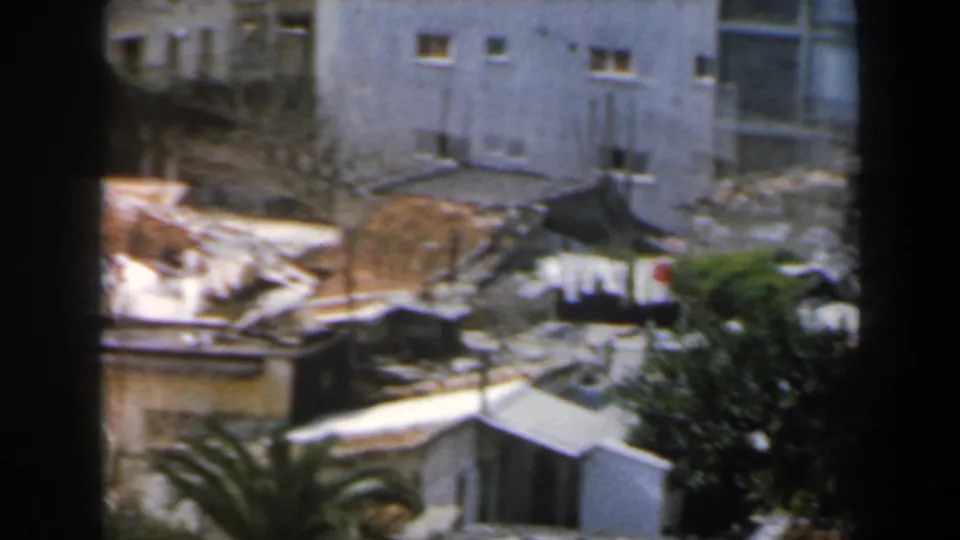 1960 8mm frame in Israel showing mixed tiled and flat roofs, pale walls, a tree canopy, and a palm frond from above.
