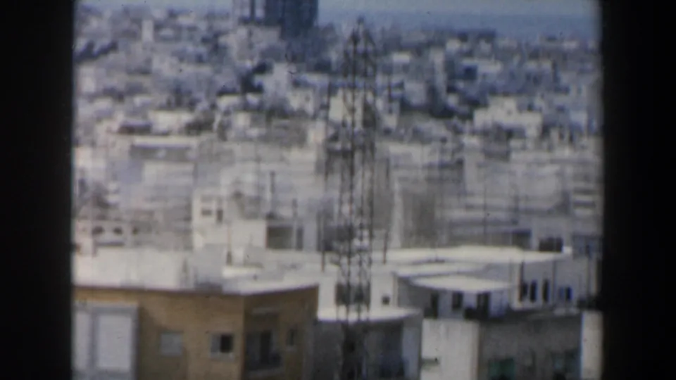 1960 Israel city view showing flat rooftops, tall antenna structures, dense low-rise buildings, and haze beyond the window frame.