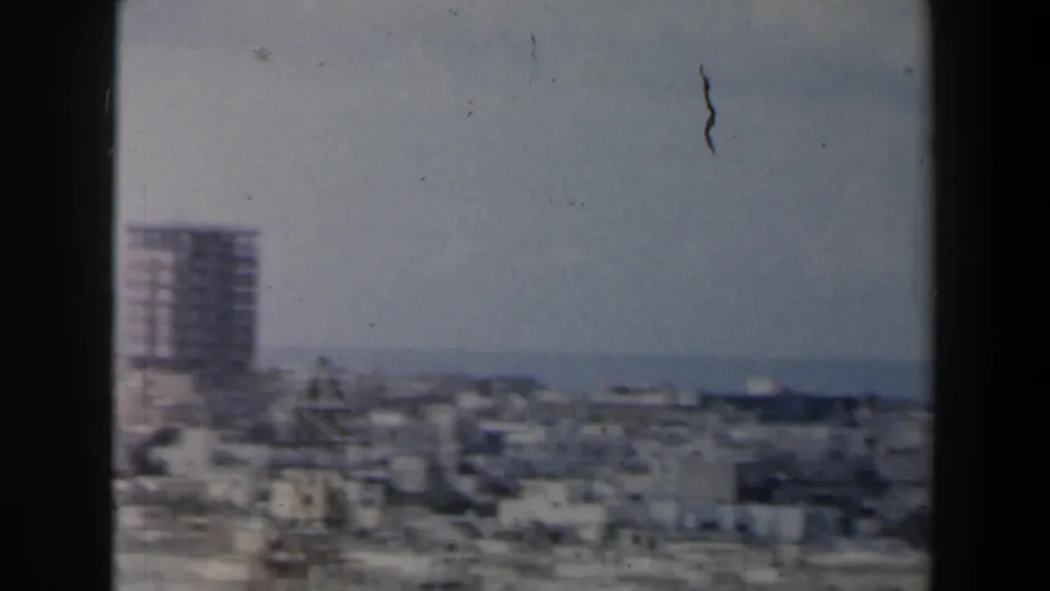 1960 Israel 8mm frame with a distant high-rise, low urban rooftops, pale sky, and possible water or hills on the horizon.