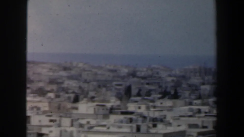 1960 Israel city panorama with low rooftops, window edge, hazy distance, and a possible body of water near the horizon.
