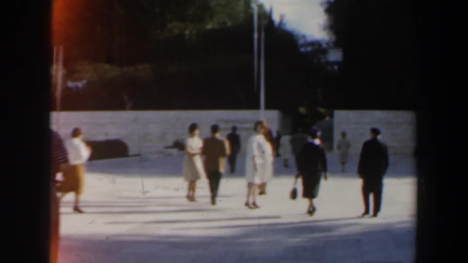 1960 Israel street-level frame showing pedestrians in period clothing walking near a low wall and trees.