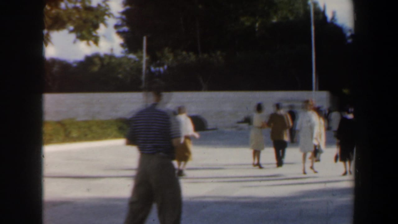1960s Israel home-movie frame showing passersby near a pale public stone wall, with a striped-shirted man in the foreground.