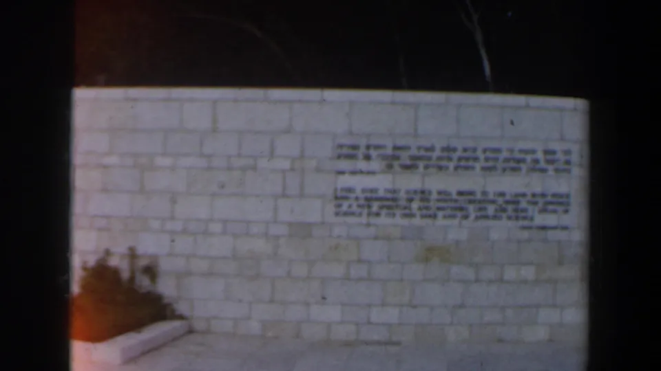 Close view of a pale stone wall in Israel with Hebrew and English inscription panels, framed by a walkway and planting bed.