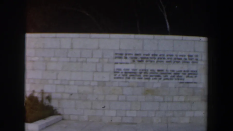 1960s Israel frame focused on a public stone wall bearing Hebrew and English text, with soft 8mm exposure and visible film grain.