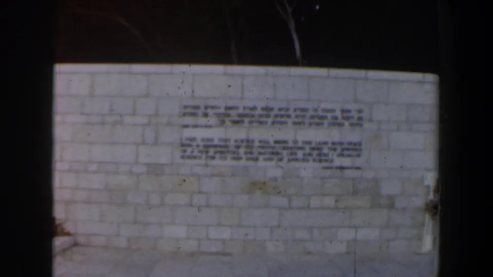 Final frame of the Israel home movie holds on the bilingual stone-wall inscription, with a partial passerby visible at the edge.