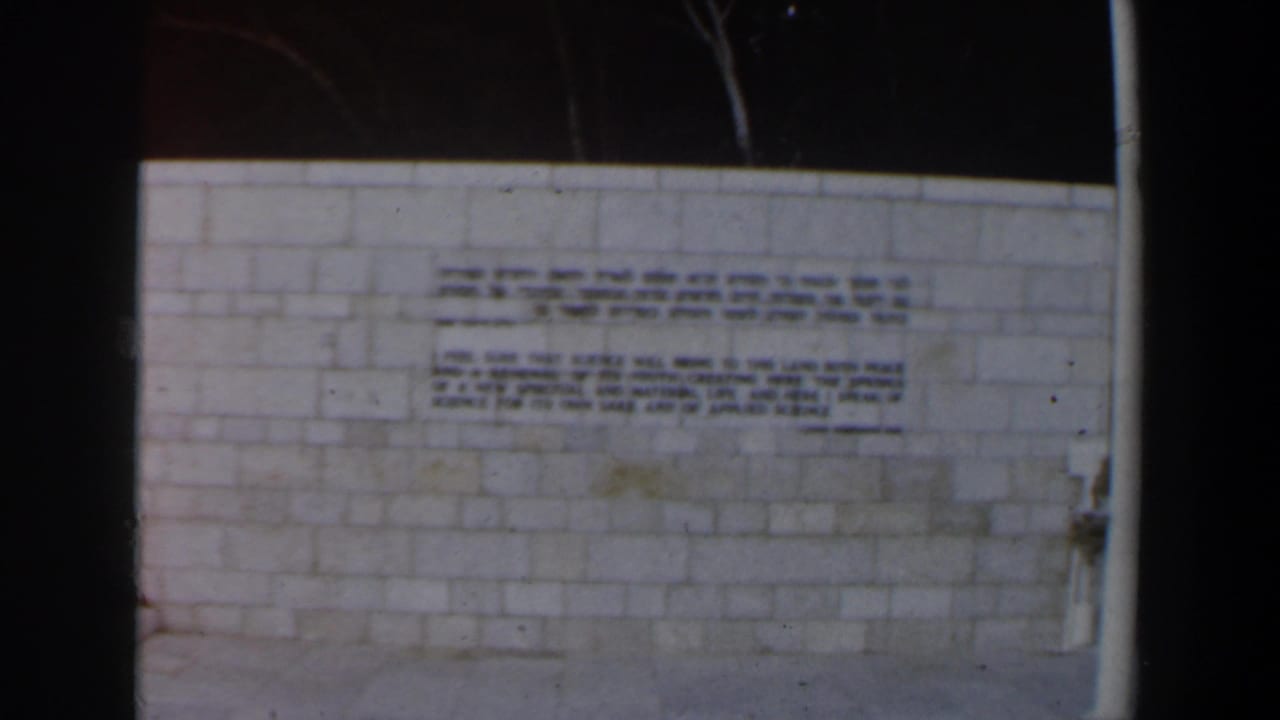Pale stone wall with dark Hebrew and English inscriptions beside a white vertical pole.