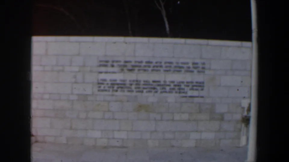 1960 Israel home-movie frame showing a pale block-stone wall with Hebrew and English inscriptions in low light.