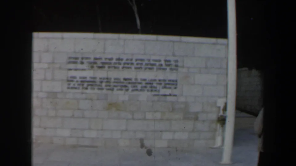 1960 Israel frame showing an inscribed pale stone wall, a white pole, and dark surrounding space in low light.