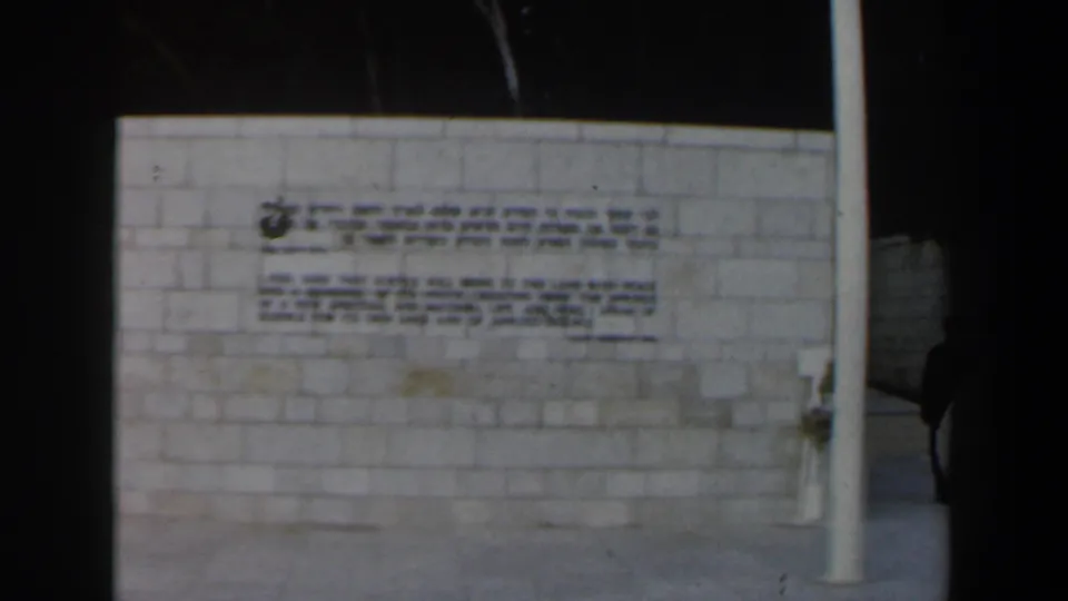 1960 Israel frame showing the inscription wall, white pole, and a partial person at the edge of the shot.