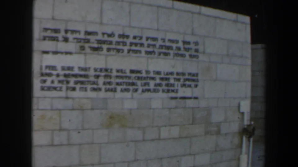 1960 Israel 8mm frame of Hebrew and English inscription on stone, with a metal pipe or fixture at the right edge.